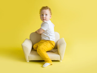 Obraz na płótnie Canvas Beautiful red-haired little toddler sitting on a beige leather sofa on a yellow background. Advertising children's furniture