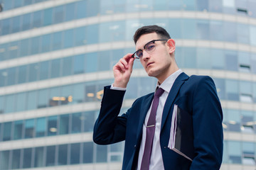 Serious self-confident bold young businessman with a file or folder for documents holding his glasses, standing outdoor office modern building or business centre, looking down at camera in formal suit