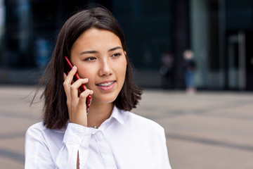 Asian young businesswoman talking on cell phone, formal dressed in white shirt, smiling outside office. Beautiful Chinese girl with braces having business conversation on smartphone outdoor work place