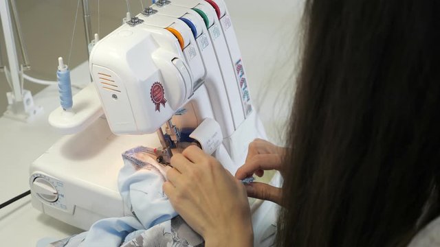 Finish detail with serger overlock stitch. Seamstress brunette woman sewing in workshop atelier on overlock cloth edges, hands closeup. Processing tissue overlock. Tailoring business in factory.