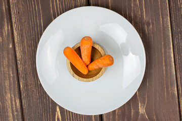 Group of three whole stale  orange stale baby carrot baby on white porcelain plate in wooden cup flatlay isolated on brown wood