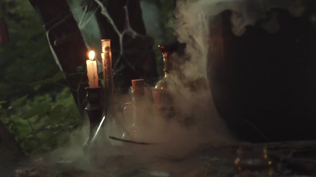 B-roll for magical video about alchemy and potions, orange warm fire from burning candle next to glass flasks on wooden table in white thick fog from cauldron, forest sorceress and fortuneteller