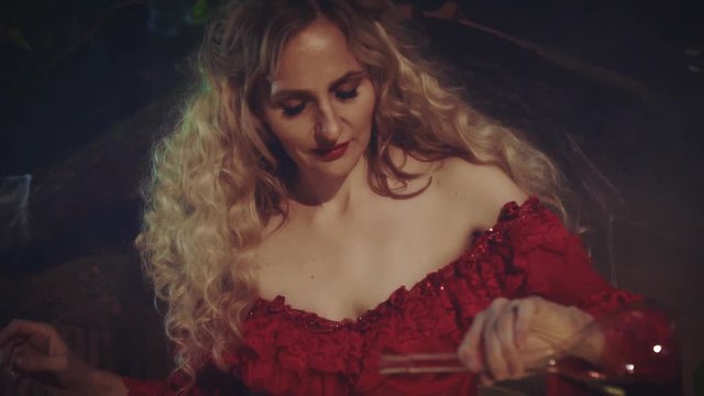 Mysterious blond woman in a red baroque dress with bare shoulders and lace. She holds a jar of red liquid in her hands and pours it into a boiling vat with steam. Enchanted night forest background