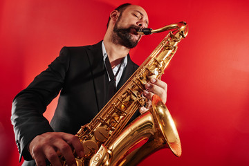 Naklejka na ściany i meble Portrait of professional musician saxophonist man in suit plays jazz music on saxophone, red background in a photo studio, bottom view