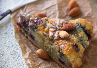 Close-up the slice of cake with berries and almonds decorated with powdered sugar on a brown paper napkin. Almonds lie next to the cut piece of cake. Homemade bakery cake. Delicious breakfast dessert.