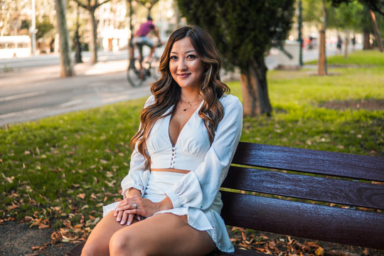 Beautiful Asian girl smiling in the park. She is wearing white dress. Happy image sitting on a bench
