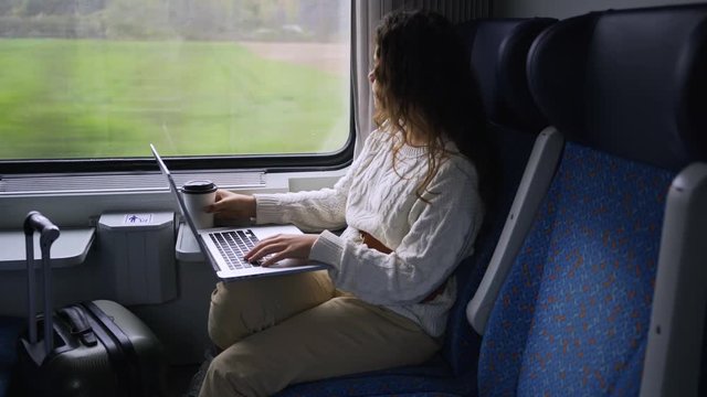 Young girl travelling on train in daytime. Woman sitting near window working on computer. View through glass females drinking coffee and enjoying landscape view passing by at background. 