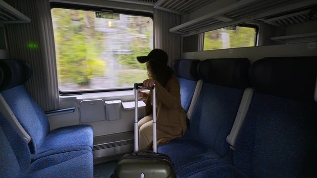 Young girl travelling on train. Wide angle view charming woman in hat and coat walking on passage inside wagon. She rolls suitcase into compartment and take a place near window