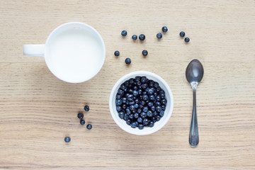 blueberry berries in a white bowl, milk in a white ceramic cup, silver spoon, berries scattered on a wooden table