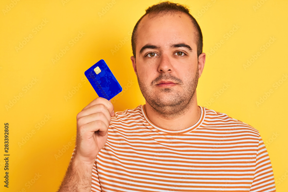 Sticker Young man holding blue credit card standing over isolated yellow background with a confident expression on smart face thinking serious