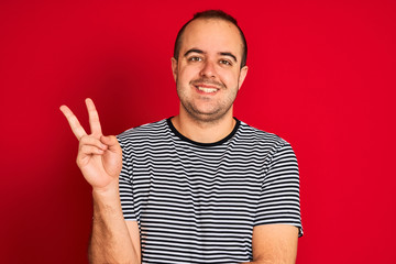Fototapeta premium Young man wearing striped navy t-shirt standing over isolated red background smiling with happy face winking at the camera doing victory sign. Number two.