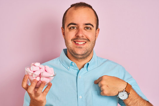 Young man holding bowl with marshmallows standing over isolated pink background with surprise face pointing finger to himself