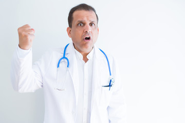 Fototapeta premium Middle age doctor man wearing stethoscope and medical coat over white background angry and mad raising fist frustrated and furious while shouting with anger. Rage and aggressive concept.