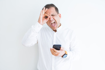 Middle age man using smartphone over white wall with happy face smiling doing ok sign with hand on eye looking through fingers