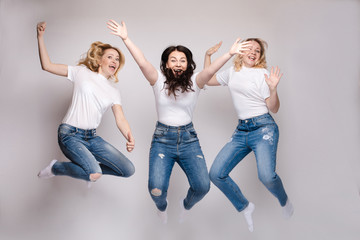 Obraz premium Close up of two blonde and one brunette women getting fun together. Three friends in casual clothes dancing and jumping. Beautiful happy girls in white t-shirts and jeans smiling and hanging out.