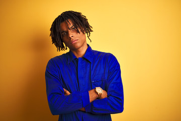 Afro worker man with dreadlocks wearing mechanic uniform over isolated yellow background skeptic and nervous, disapproving expression on face with crossed arms. Negative person.