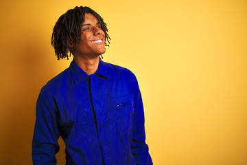 Obraz premium Afro worker man with dreadlocks wearing mechanic uniform over isolated yellow background looking away to side with smile on face, natural expression. Laughing confident.