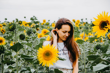 A beautiful young woman with long hair is standing in a field of sunflowers looking down