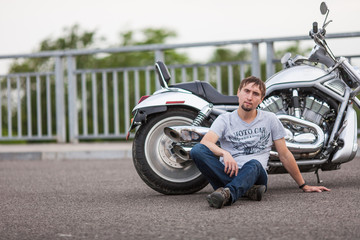 Handsome rider man sitting next to modern powerful shiny motorcycle on an open road. Brutal young man sitting at powerful shiny style cruiser motorbike on highway