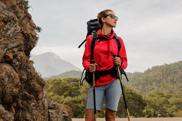 Fototapeta na wymiar Girl in sunglasses standing on the rocks with hiking backpack and walking sticks