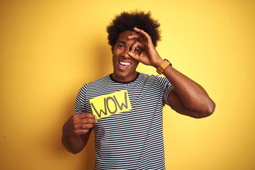 American man with afro hair holding wow banner standing over isolated yellow background with happy face smiling doing ok sign with hand on eye looking through fingers