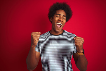 Young american man with afro hair wearing navy striped t-shirt over isolated red background very happy and excited doing winner gesture with arms raised, smiling and screaming for success.