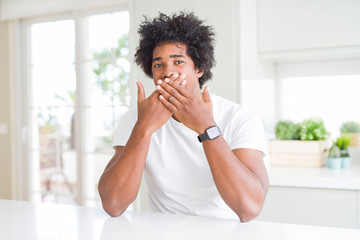 Fototapeta na wymiar Young african american man wearing casual white t-shirt sitting at home laughing and embarrassed giggle covering mouth with hands, gossip and scandal concept
