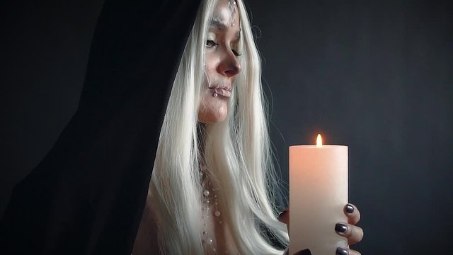 mystical video in Gothic style, woman with blond hair with sweet white skull and black cloak holding burning candle in hands, image for masquerade for Halloween and Day of Dead, lady blows out fire