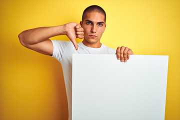 Young handsome man holding advertising banner over yellow isolated background with angry face,...