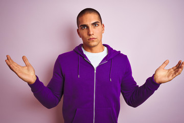 Young handsome man wearing purple sweatshirt over pink isolated background clueless and confused expression with arms and hands raised. Doubt concept.