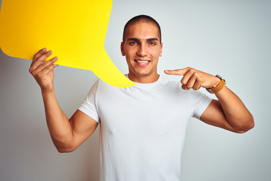 Young man holding yellow speech bubble over white isolated background very happy pointing with hand and finger