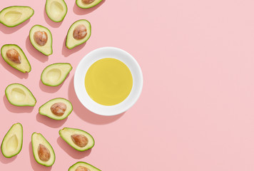 Avocado oil in a bowl and avocado halves on pink background, flat lay with copy space