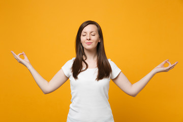 Relaxed young woman in white casual clothes keeping eyes closed hold hands in yoga gesture, relaxing meditating isolated on yellow orange wall background. People lifestyle concept. Mock up copy space.