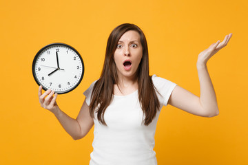 Portrait of shocked young woman in white casual clothes spreading hands, holding round clock...