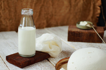 A glass bottle with cow's milk close-up stands on a wooden plank, gauze lies nearby. In the foreground and background cottage cheese is homemade.
