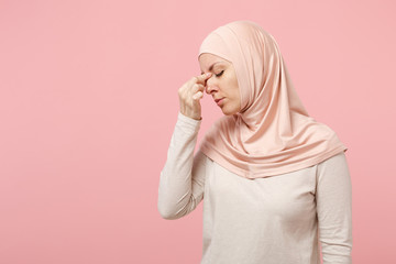 Tired arabian muslim woman in hijab light clothes posing isolated on pink background in studio. People religious Islam lifestyle concept. Mock up copy space. Keeping eyes closed, putting hand on nose.