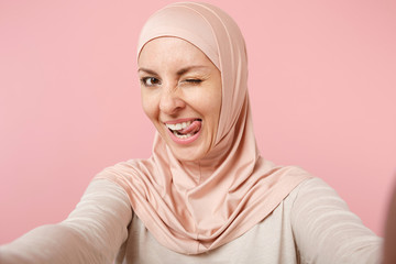 Close up arabian muslim woman in hijab light clothes posing isolated on pink background. People religious Islam lifestyle concept. Mock up copy space. Doing selfie shot on mobile phone showing tongue.