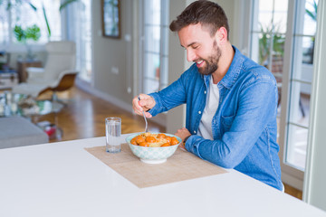 Handsome man eating pasta with meatballs and tomato sauce at home while smiling at the camera