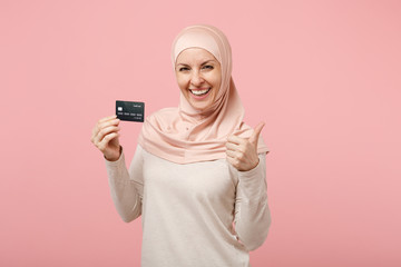 Cheerful young arabian muslim woman in hijab light clothes posing isolated on pink background. People religious Islam lifestyle concept. Mock up copy space. Holding credit bank card, showing thumb up.
