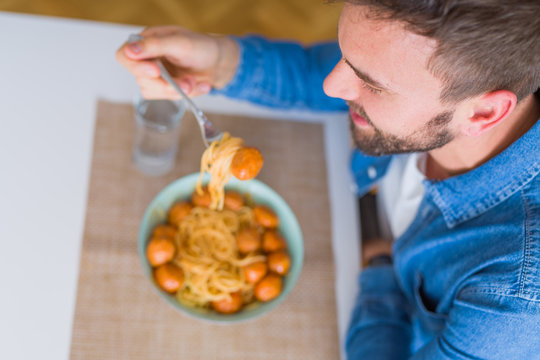 Handsome man eating pasta with meatballs and tomato sauce at home while smiling at the camera