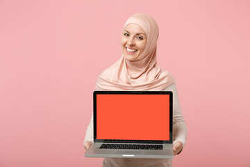 Smiling arabian muslim woman in hijab light clothes posing isolated on pink background. People religious Islam lifestyle concept. Mock up copy space. Hold laptop pc computer with blank empty screen.