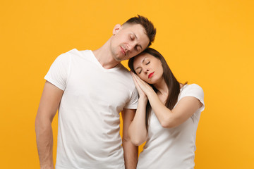 Young couple two friends guy girl in white empty blank design t-shirts posing isolated on yellow orange background. People lifestyle concept. Mock up copy space. Sleep with folded hands under cheek.