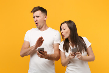 Shocked young couple friends guy girl in white empty blank t-shirts posing isolated on yellow orange background. People lifestyle concept. Mock up copy space. Using mobile phone, typing sms message.