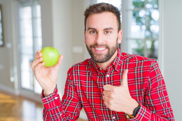 Handsome man eating fresh healthy green apple happy with big smile doing ok sign, thumb up with fingers, excellent sign