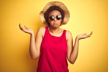 African american woman wearing summer hat and sunglasses over yellow isolated background clueless...