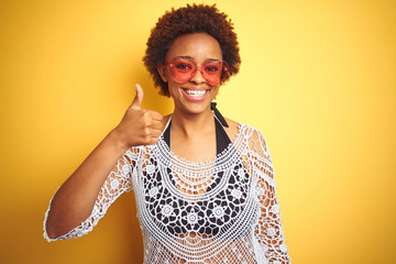 Young african american woman with afro hair wearing bikini and heart shaped sunglasses doing happy thumbs up gesture with hand. Approving expression looking at the camera with showing success.