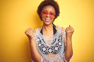 Young african american woman with afro hair wearing bikini and heart shaped sunglasses celebrating surprised and amazed for success with arms raised and open eyes. Winner concept.
