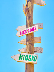 Tall post with colored wooden signboards pointing to various destinations and a beach services in Spanish on a blue sky. Kiosk and Ice Creams.
