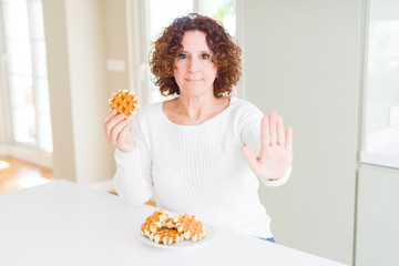 Senior woman eating sweet belgian waffle with open hand doing stop sign with serious and confident expression, defense gesture