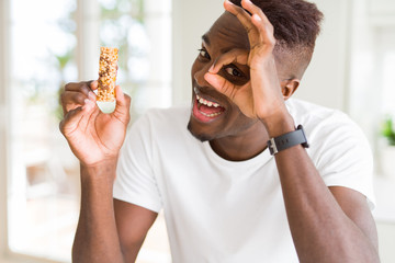 African american man eating energetic cereals bar with happy face smiling doing ok sign with hand...